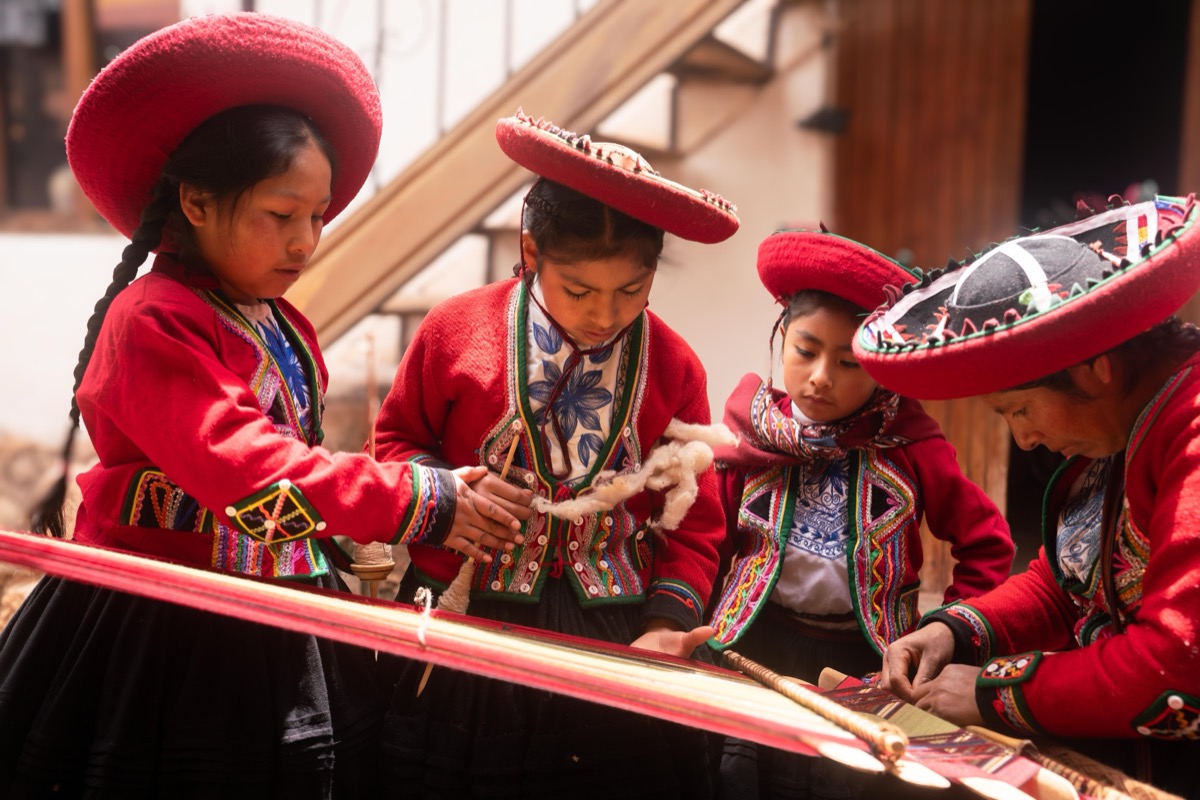 Four young girls in traditional Chinchero red dress and hats learning to weave at a loom, Cusco, Peru