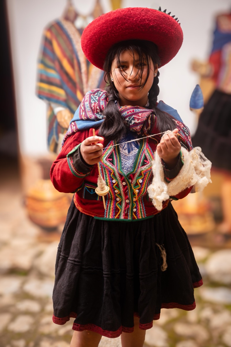 Young girl in traditional Chinchero dress and red hat spinning wool with a drop spindle, Cusco, Peru