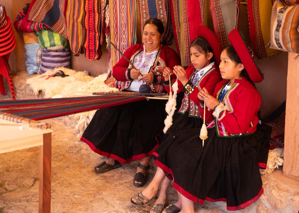 A Colores de Abundancia weaver teaching two young girls traditional loom weaving in traditional Chinchero dress, Cusco, Peru