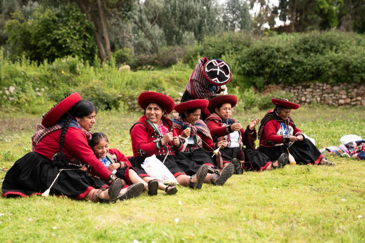 A group of women and girls in traditional Chinchero dress sitting on the grass spinning wool together outdoors, Cusco, Peru