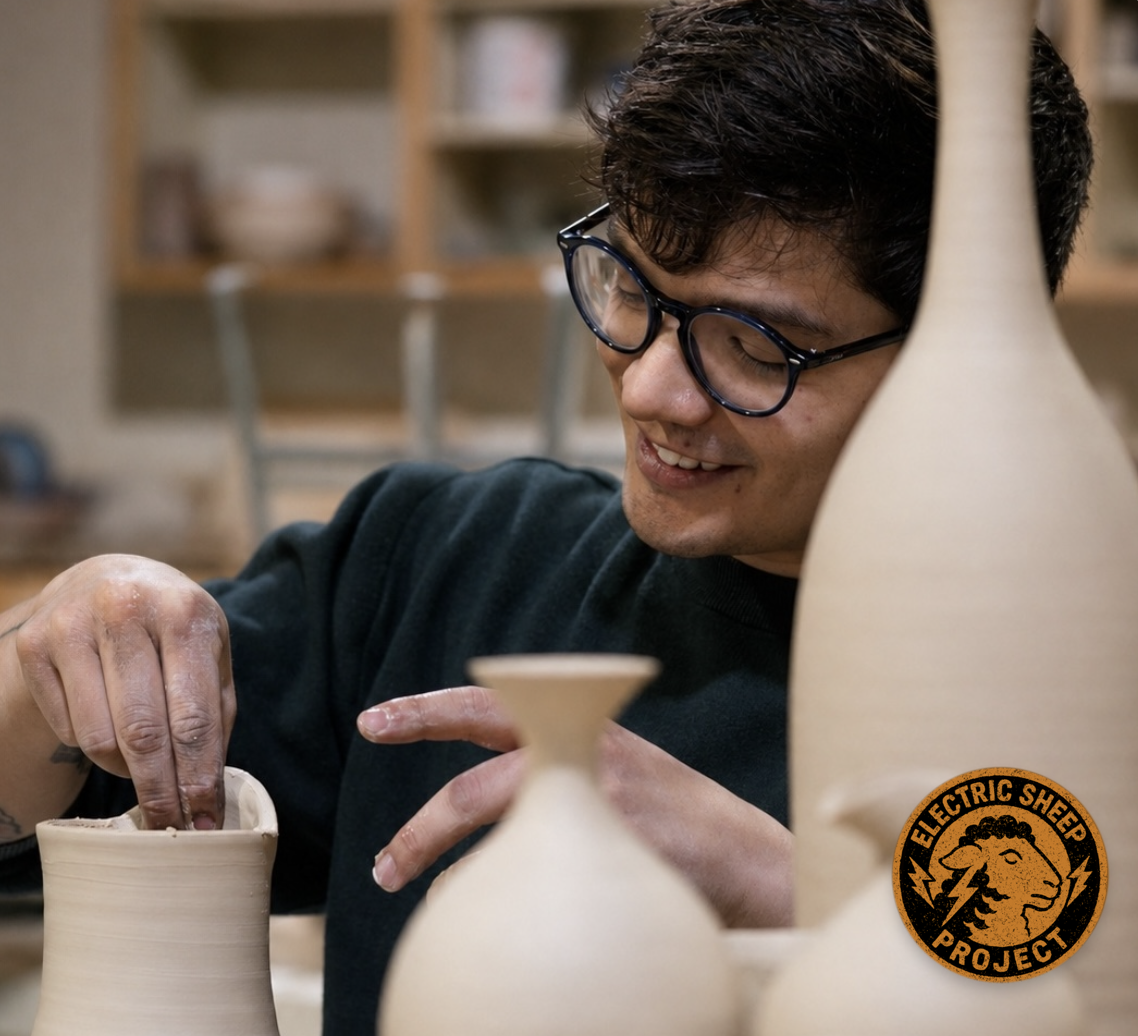 Young man with glasses shaping clay on a pottery wheel surrounded by ceramic vases at the Electric Sheep Project studio