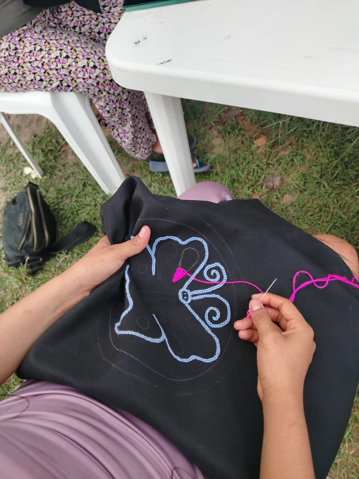 Hands of Ida Castro Rojas embroidering kené design in pink thread on black fabric outdoors, Paohyan, Loreto, Peru