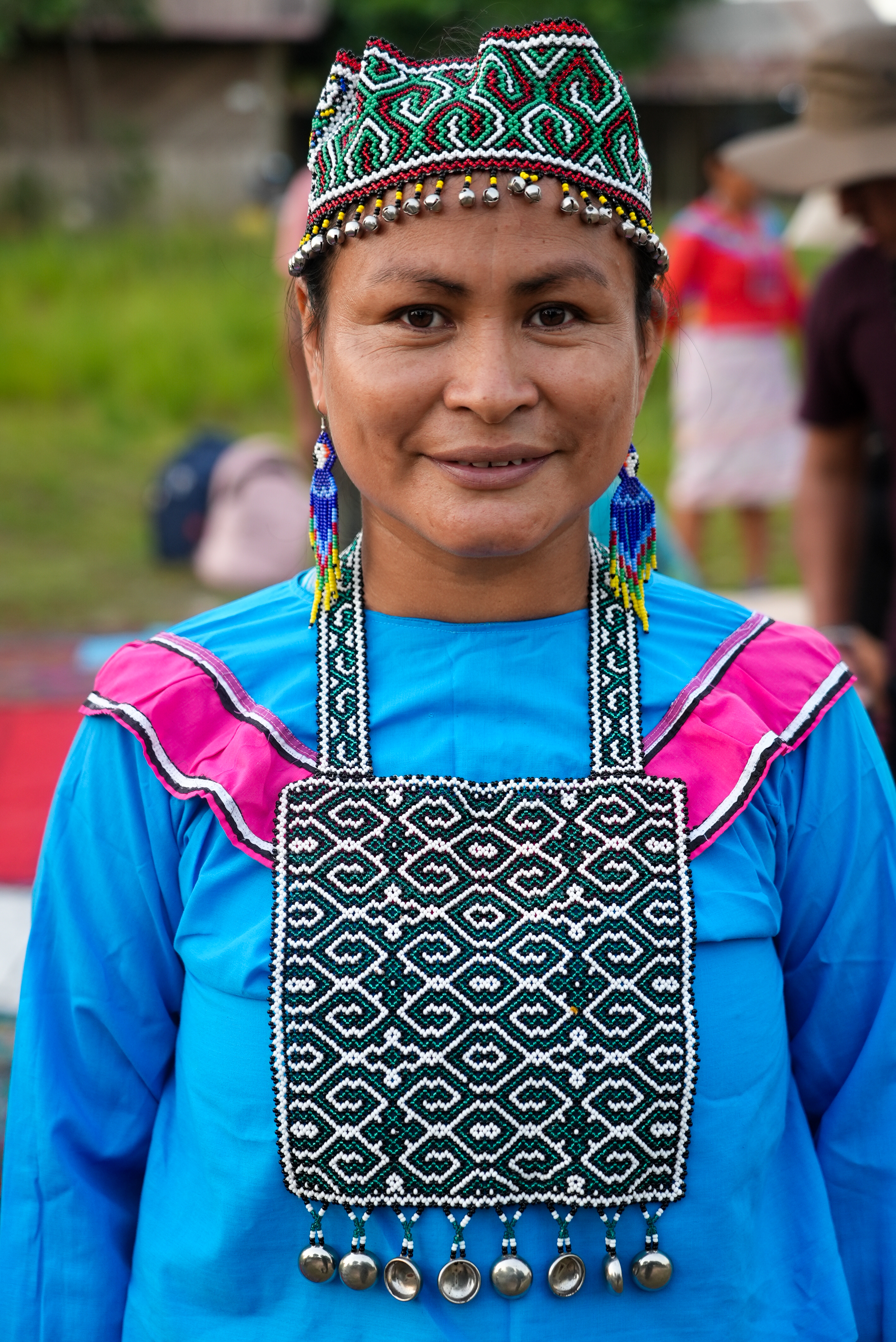Shipibo-Konibo woman in traditional blue dress wearing intricate kené beadwork breastplate, earrings, and beaded crown with bells, Peru