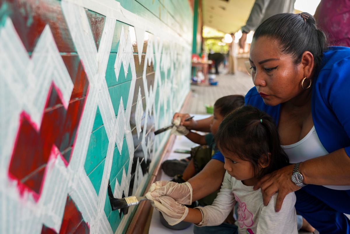 Woman guiding a young child's hand as they paint a geometric kené-patterned mural together, Peru