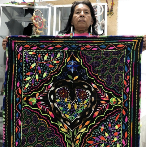 Olga Morí Diaz holding up a large kené textile with vibrant geometric designs on black fabric, Calleria, Pucallpa, Peru