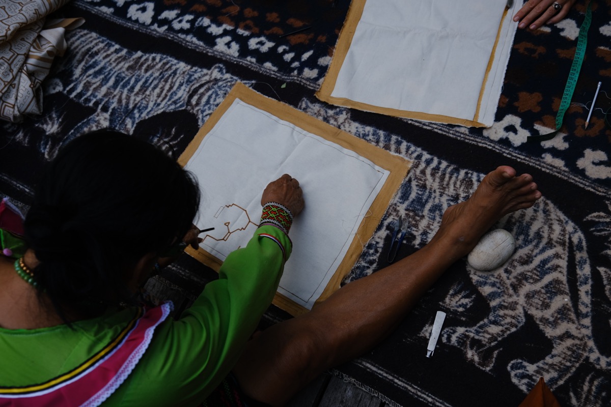 Shipibo-Konibo artist drawing kené design onto white canvas surrounded by patterned textiles, Calleria, Pucallpa, Peru