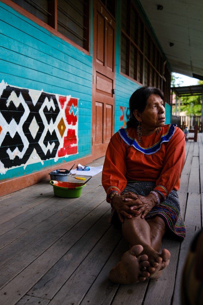 Olga Morí Diaz seated on a wooden porch in traditional Shipibo-Konibo dress before a turquoise wall painted with kené murals, Comunidad Nativa de Calleria, Pucallpa, Peru