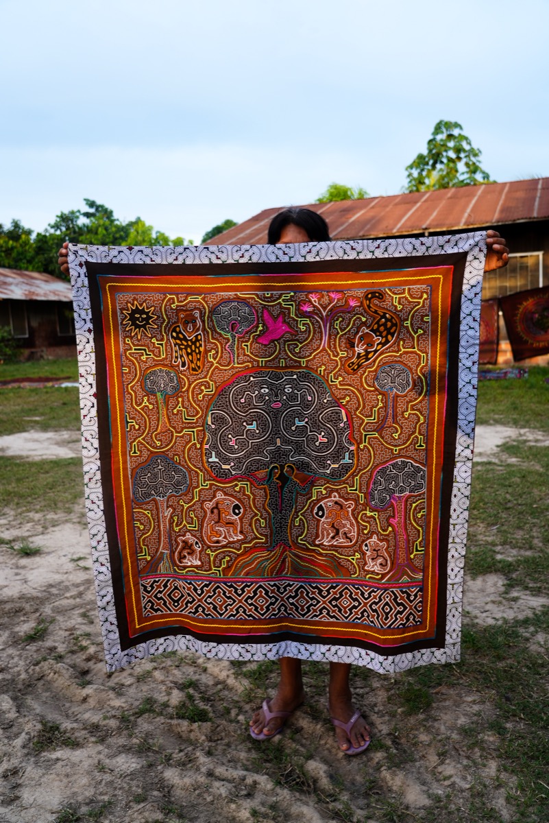 Shipibo-Konibo artisan holding a large kené tela pintada featuring a tree of life with animals and geometric patterns, Calleria, Pucallpa, Peru