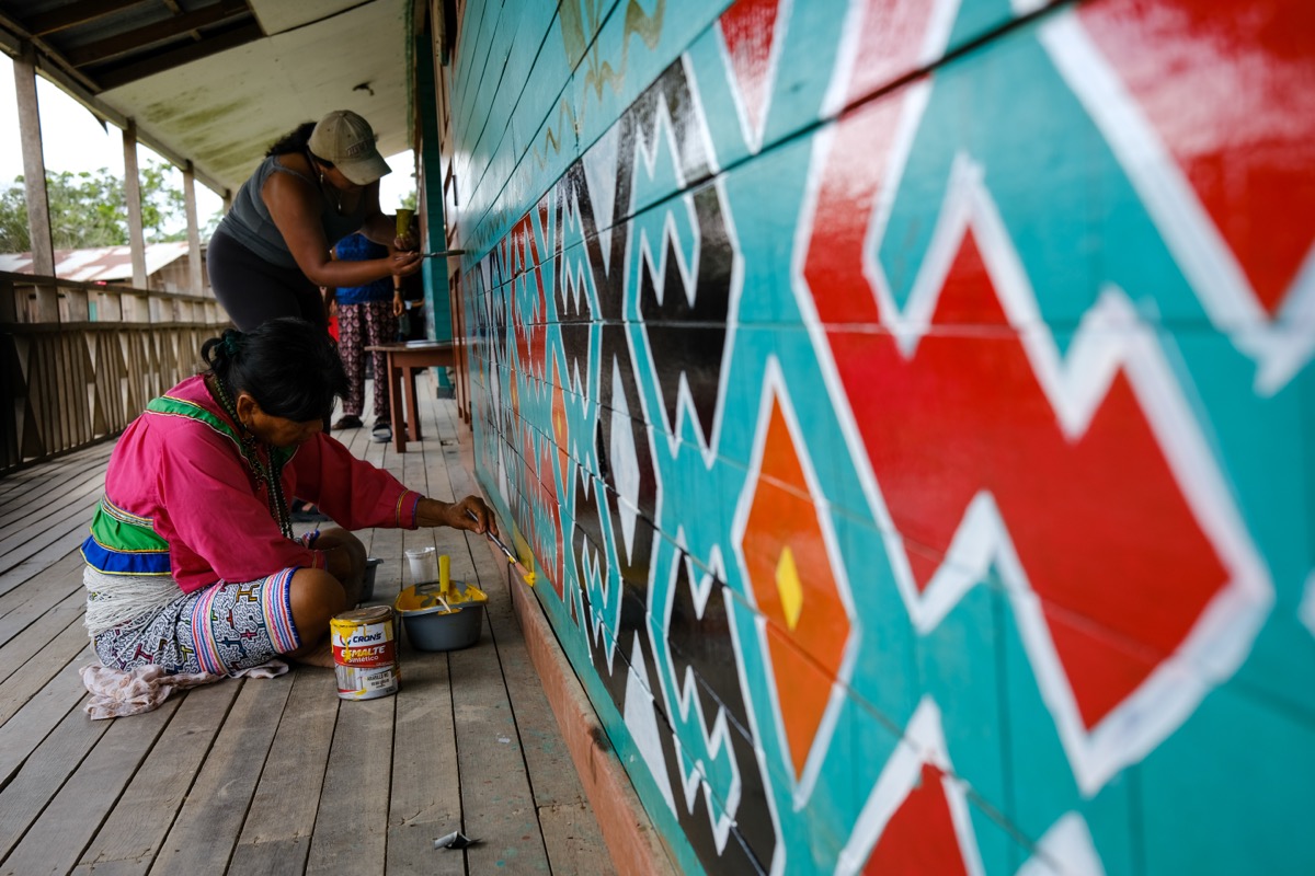 Shipibo-Konibo artists painting a large kené mural in turquoise, red, and orange on a wooden wall in Calleria, Pucallpa, Peru