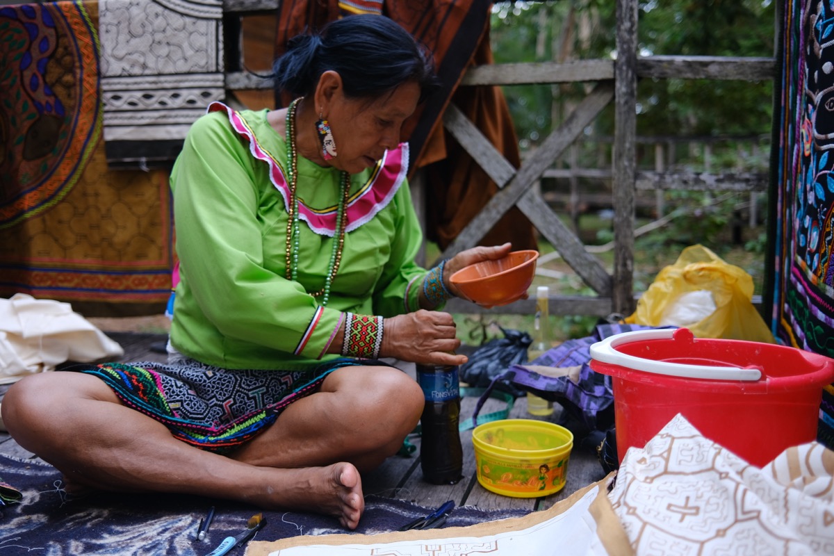 Olga Morí Diaz seated cross-legged mixing natural pigments while painting kené designs, with finished textiles hanging behind her, Pucallpa, Peru