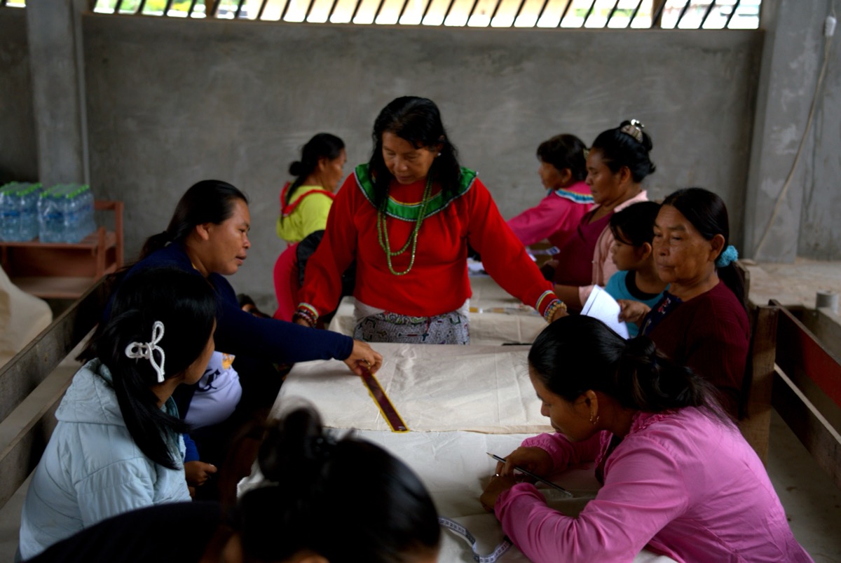 Olga Morí Diaz in red blouse and beaded necklace guiding Shipibo-Konibo women through a kené textile workshop in Calleria, Pucallpa, Peru