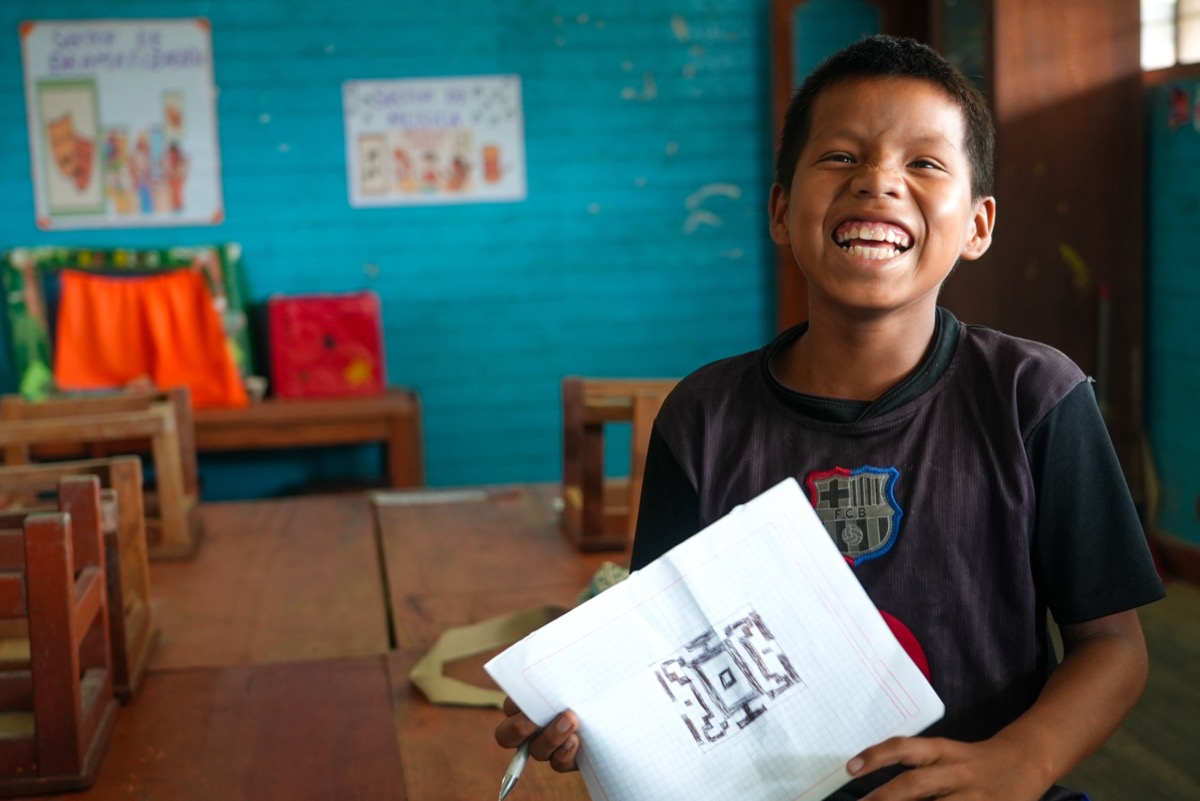 Young student laughing joyfully in a classroom while holding a notebook with a hand-drawn design, Peru